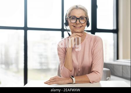 Ritratto di una bella donna bionda matura sorridente o rappresentante del servizio clienti di media età seduto alla reception contro panoramica finestra Foto Stock