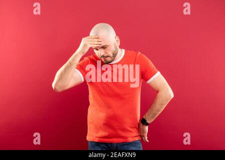 Un uomo calvo di mezza età con barba e una T-shirt rossa su sfondo rosso. Tiene la testa con le mani. Ha un mal di testa. Foto Stock