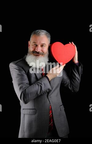 Mature uomo bearded abito grigio tiene rosso cuore forma scatola regalo, isolato sfondo nero Foto Stock