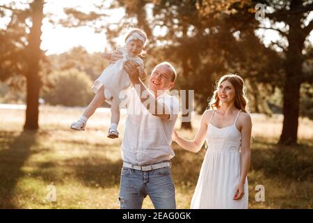 Ritratto di una madre, padre tiene, getta e gira la figlia sulle mani che camminano sulla natura durante la vacanza estiva. Mamma, papà e ragazza che giocano in t Foto Stock