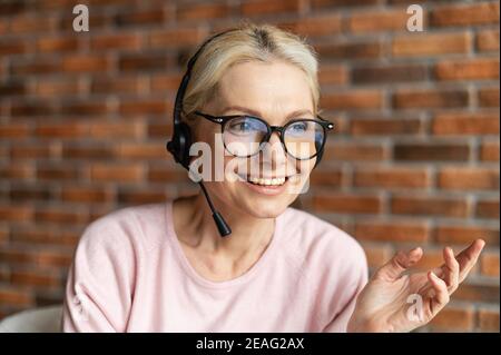 Primo piano del volto di un'affascinante signora bionda matura ed elegante, indossando cuffie, occhiali, sorridendo e guardando lontano. Ritratto di mezza età Foto Stock