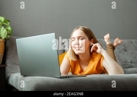 Felice relax giovane donna si trova sul divano con computer portatile in cuffie. Lavoro a distanza, studio online, shopping online guardando film o videochiamata su computer portatile Foto Stock