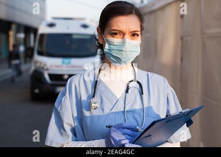 Giovane donna NHS UK EMS medico di fronte alla sanità Struttura ICU, indossare una maschera protettiva per il viso che tiene il controllo medico delle condizioni del paziente Forma, Coronavirus Foto Stock