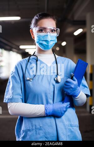 Ritratto di giovane bella donna medico che indossa uniforme blu, occhiali di sicurezza & maschera facciale, preoccupato, overlavored & stressed, UK NHS ICU chiave frontline medica Foto Stock