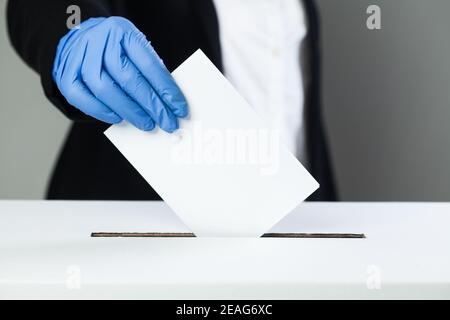 Persona in tuta nera che indossa guanti di gomma di lattice protettivo blu mettendo carta da voto nella casella di voto, giorno delle elezioni durante Coronavirus COVID-19 pandemic c Foto Stock