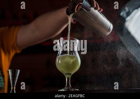 Il barista spruzzi su vetro illuminato con un cocktail freddo verde brillante sul bancone del bar e fa fuoco su di esso. Foto Stock