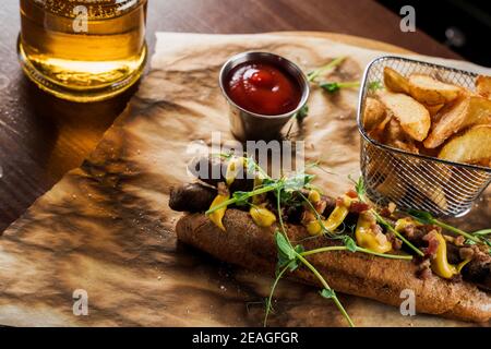 Barbecue hot dog alla griglia e birra con senape gialla e ketchup su sfondo di legno. Sandwich hot dog con patatine fritte e salse. Foto Stock