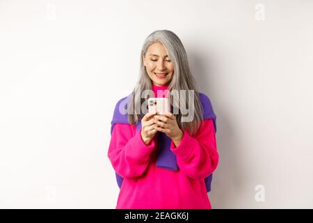 Bella donna asiatica anziana in rosa elegante maglione utilizzando smartphone. Vecchia signora che legge sul telefono cellulare e sorridente, in piedi su sfondo bianco Foto Stock