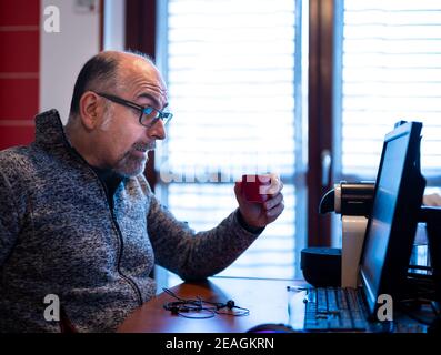 Un uomo caucasico di mezza età che lavora da casa, mentre beve un caffè, è piacevolmente sorpreso da una notifica che legge sul suo monitor del computer. Foto Stock