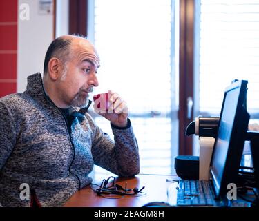 Un uomo caucasico di mezza età che lavora da casa, mentre beve un caffè, è piacevolmente sorpreso da una notifica che legge sul suo monitor del computer. Foto Stock