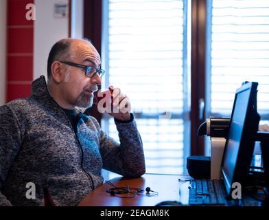 Un uomo caucasico di mezza età che lavora da casa, mentre beve un caffè, è piacevolmente sorpreso da una notifica che legge sul suo monitor del computer. Foto Stock