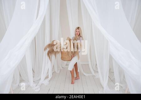 Giovane bionda donna in una camicia lilla, vestito bianco, tiene nelle sue mani paglia cesto con pampas erba tra i tessuti ariosi e tulle sviluppo. Minimali Foto Stock