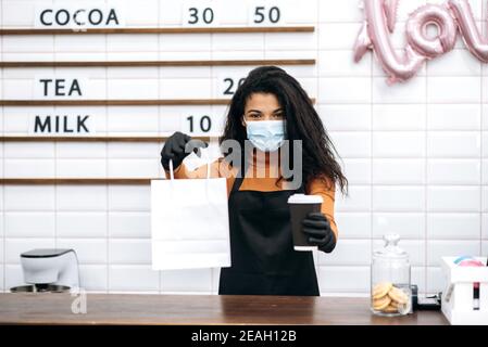 Giovane barista afroamericano ragazza in uniforme e maschera medica e guanti protettivi dà un caffè visitatore in un bicchiere di cartone monouso e dessert in una borsa di carta per andare. Concetto di caffè in movimento. Foto Stock