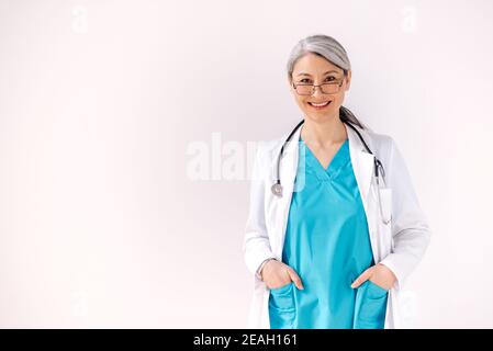 Medico caucasico sicuro in uniforme medica e stetoscopio si trova su uno sfondo bianco, guarda la macchina fotografica e sorride. Concetto di assistenza sanitaria Foto Stock