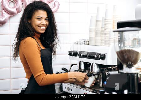 Vista laterale una sorridente e amichevole barista afroamericana fa caffè in tazza di carta per i visitatori del caffè utilizzando la macchina del caffè, in piedi dietro il bancone del bar e sorridente Foto Stock