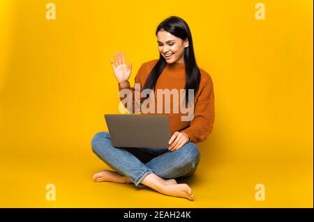 Attraente bruna caucasica ragazza si leva su uno sfondo arancione isolato tiene un laptop guarda direttamente nella fotocamera con sorriso amichevole, seduta su isolato sfondo arancione Foto Stock