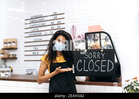 Triste cameriera afroamericana, barista o piccola impresa proprietario. Giovane donna infelice in un grembiule nero e maschera medica, si trova all'interno di una caffetteria, un ristorante o un bar e contiene un cartello chiuso Foto Stock