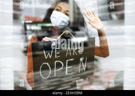 African American piccolo imprenditore in maschera medica tiene un cartello SIAMO APERTI all'ingresso di un ristorante, bar, caffetteria o negozio, e in attesa di visitatori Foto Stock