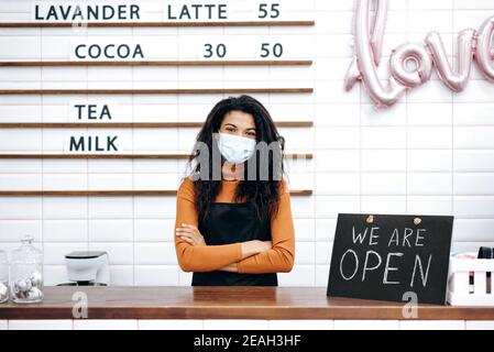 Ritratto di barista o cameriera afro-americana che indossa una maschera medica, si trova dietro il banco bar accanto a un cartello SIAMO APERTI, in attesa di visitatori Foto Stock