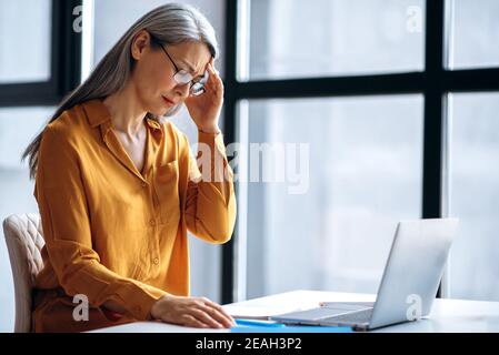 Triste asiatico medio invecchiato grigio-capelli donna d'affari o tutor femminile, è stanco di lavoro straordinario on-line, o di condurre insegnamento a distanza, chiuso gli occhi, mal di testa, bisogno di un riposo Foto Stock