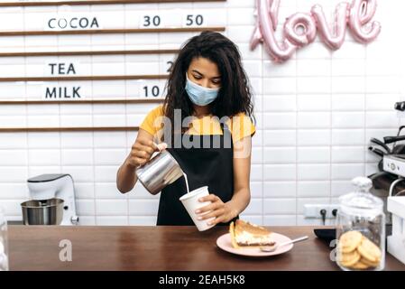 Happy african american barista femminile o proprietario di una caffetteria in grembiule nero e maschera medica prepara il caffè mentre si trova al bancone del bar, versa il latte in un bicchiere di caffè di cartone usa e getta Foto Stock