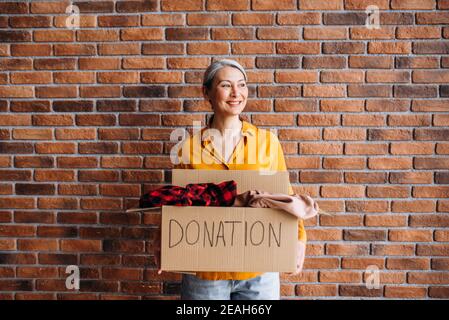 La volontaria femminile asiatica con capelli grigi senior contiene una scatola di cartone con una donazione, per il riutilizzo o il riciclaggio di vecchi abiti. Ritratto di una donna felice si trova contro muro di fondo in mattoni con scatola di donazione Foto Stock