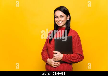 Bella bruna caucasica ragazza si trova su uno sfondo arancione isolato tiene un laptop guarda direttamente nella fotocamera con amichevole sorridi Foto Stock