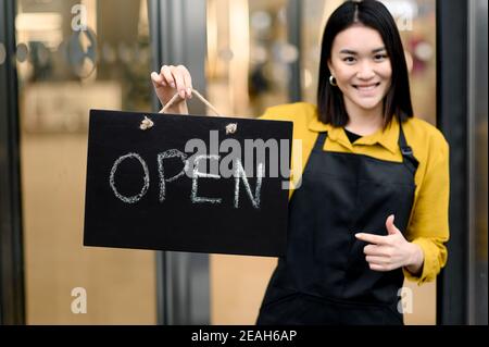 Benvenuto, siamo aperti. Una giovane donna, cameriera o proprietario di un bar o di un ristorante che indossa un grembiule nero si erge all'ingresso con un cartello APERTO e sorridente. Piccola impresa Foto Stock