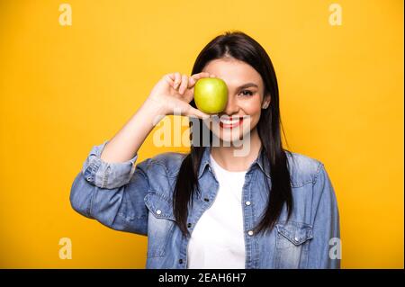 Donna caucasica felice conduce uno stile di vita sano. Una ragazza si leva in piedi su uno sfondo arancione isolato, tiene una mela verde fresco, guarda nella fotocamera copre un occhio con una mela e sorrisi. Cibo sano Foto Stock