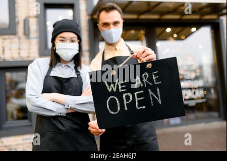 Cartello APERTO in primo piano. Due camerieri sfocati che indossano maschere protettive, uomo caucasico e donna asiatica in piedi fuori di un ristorante, bar o bar e mostrando cartello APERTO, benvenuto Foto Stock