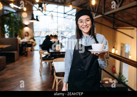 Bella donna asiatica felice cameriera o barista con occhiali si trova all'interno di un ristorante, caffè o bar, tiene una tazza di caffè aromatico e con sorriso amichevole guarda la macchina fotografica Foto Stock