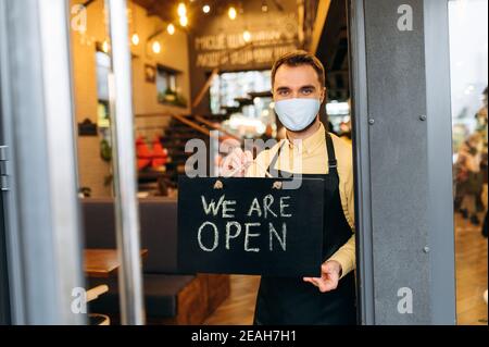 Siamo APERTI. Ritratto di un cameriere o barista caucasico soddisfatto in piedi al ristorante o all'ingresso del caffè indossando maschera medica sul viso e grembiule nero. Uomo dipendente che mostra un cartello APERTO nel suo Foto Stock