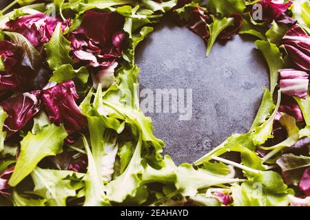 Forma del cuore da foglie di insalata verde e viola mescolano lo sfondo. Tavolo in cemento Foto Stock
