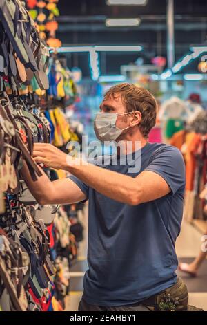 Serious uomo blu in t-shirt blu scelta scarpe in negozio, indossare maschera blu viso proteggere da virus COVID, pubblica misura di controllo della salute personale Foto Stock