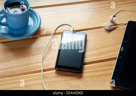 Vista dall'alto di una scrivania rustica in legno con telefono cellulare nero, tablet, auricolari e tazza di caffè. Concetto di spazio di lavoro, ufficio domestico, luogo di studio o rela Foto Stock