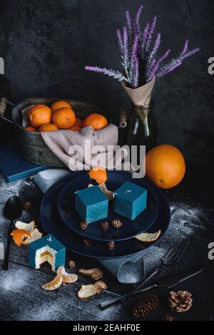 Torta blu ripiena di arancio e mandarino a forma di cubo. Vicino su un tavolo nero sono ingredienti arance, mandarini, noci. Stile rustico. Foto Stock