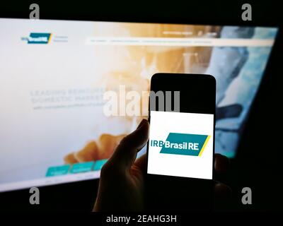 Persona che detiene il telefono cellulare con il logo della compagnia di riassicurazione IRB Brasil RE sullo schermo davanti al sito web aziendale. Focus sul display del cellulare. Foto Stock