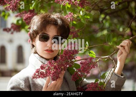 Ritratto di una giovane donna in occhiali da sole vicino a un lilla bush in un cappotto grigio in una giornata di primavera soleggiata Foto Stock