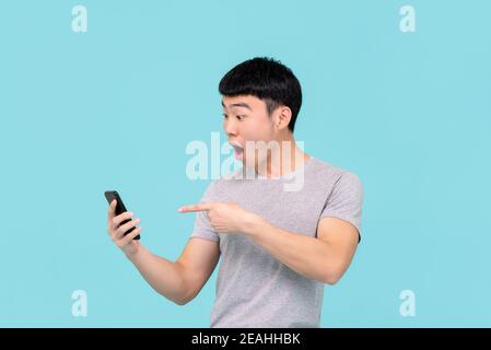 Scioccato eccitato giovane asiatico che anelava e punta al mobile telefono isolato su sfondo azzurro Foto Stock
