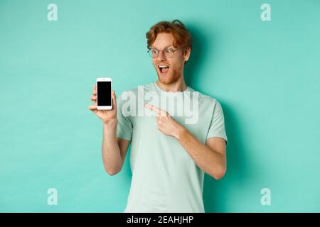 Dai un'occhiata. Bel ragazzo rosso in occhiali che punta il dito sullo schermo vuoto dello smartphone, mostrando la promozione online, in piedi stupito sopra il turchese Foto Stock