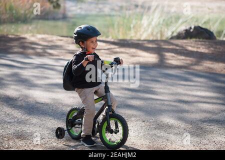 Un ragazzo di cinque anni imparando a fare un giro in bicicletta con ruote da allenamento e un lago sullo sfondo. Foto Stock