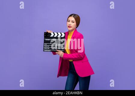 Giovane bella donna asiatica modello in abiti colorati che tengono il film clapperboard isolato su sfondo viola Foto Stock