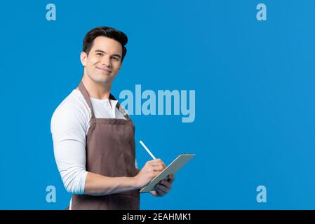 Sorridente barista giovane e bello che indossa grembiule per scrivere sulla clipboard isolato su sfondo blu Foto Stock