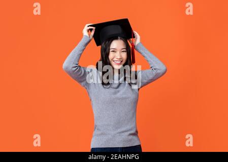 Felice sorridente giovane donna asiatica con cappuccio di graduazione isolato sopra sfondo arancione per i concetti di istruzione Foto Stock