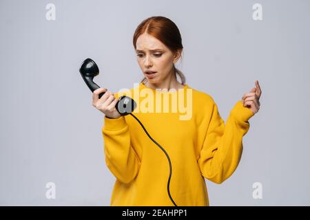 Donna giovane disperata in maglione giallo che tiene ricevitore del ricevitore del ricevitore di telefono retro e guardandolo Foto Stock