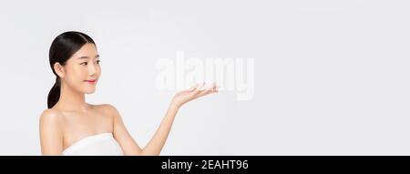 Giovane bella donna asiatica con un aspetto fresco e pulito sorridente e. guardando lateralmente a mano aperta vuota isolata su striscione bianco sfondo per la bellezza Foto Stock