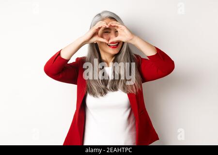 Bella donna asiatica di mezza età con capelli grigi, indossando blazer rosso, mostrando il segno del cuore e picchiando su di esso, ti amo gesto, in piedi su bianco Foto Stock