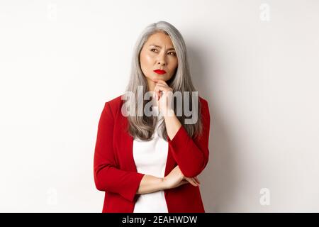 Concetto aziendale. Donna d'affari asiatica anziana premurosa che guarda da parte, pensando mentre si sta in piedi su sfondo bianco Foto Stock