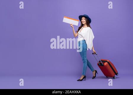 Giovane ragazza turistica asiatica sorridente con bagaglio che mostra imbarco aereo passa e cammina su uno sfondo viola colorato Foto Stock