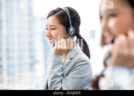 Sorridente bella donna asiatica che lavora nel call center ufficio della città in qualità di operatore del servizio clienti con team Foto Stock